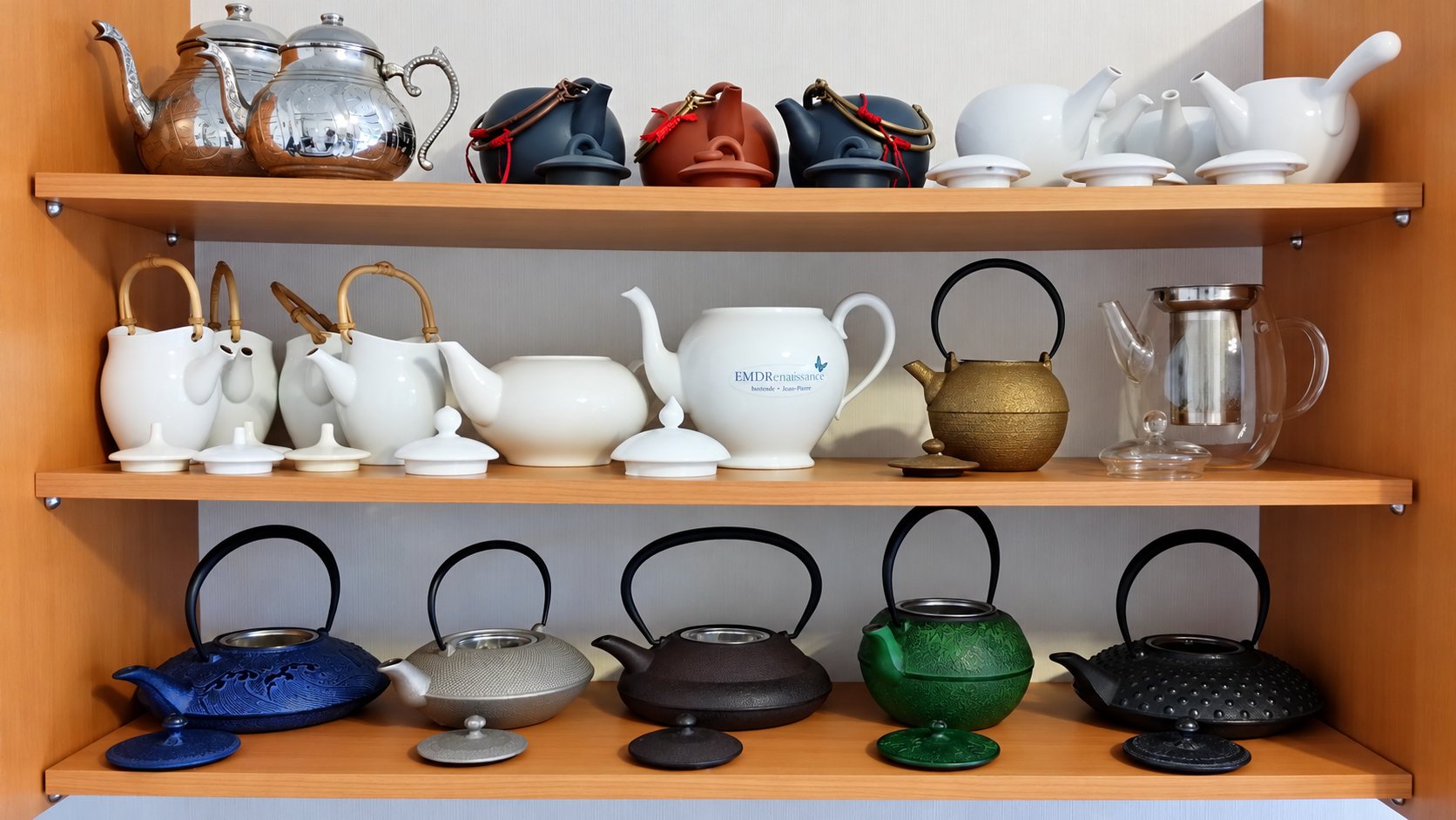 Trois étagères en bois présentant une collection de théières en métal argenté, en grès, en porcelaine blanche, en verre et en fonte de plusieurs couleurs, avec leurs couvercles.