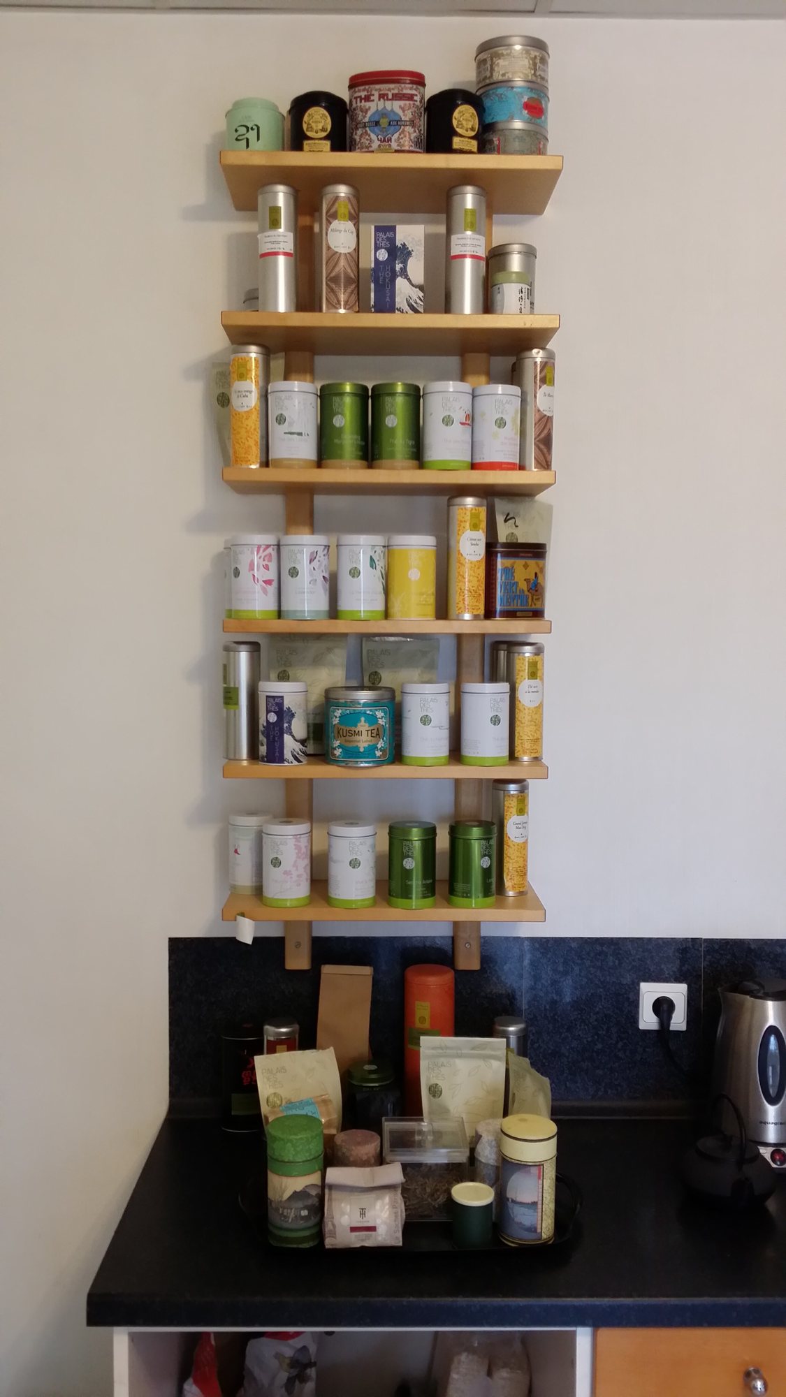 Une étagère murale en bois à cinq niveaux présente de nombreuses boîtes de thé métalliques de différentes couleurs, au-dessus d'un plan de travail noir où sont posés des sachets de thé et une bouilloire.