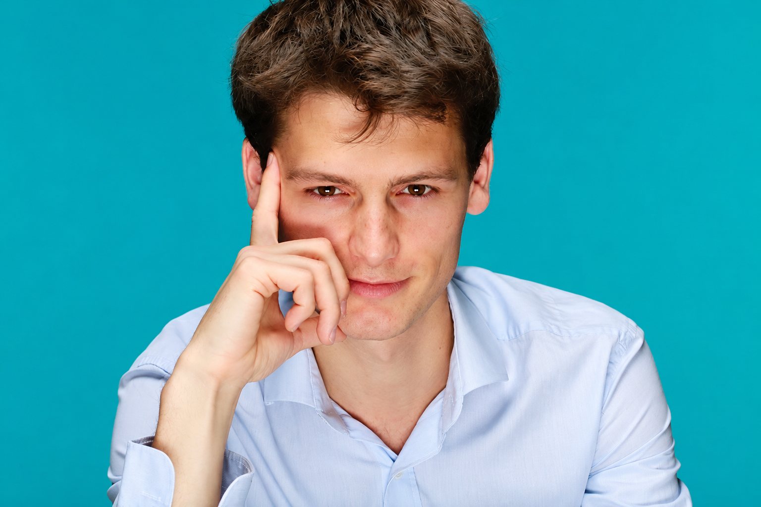 Un jeune homme aux cheveux bruns, vêtu d'une chemise bleu clair, pose la main sur la tempe avec un regard pensif devant un fond turquoise uni.