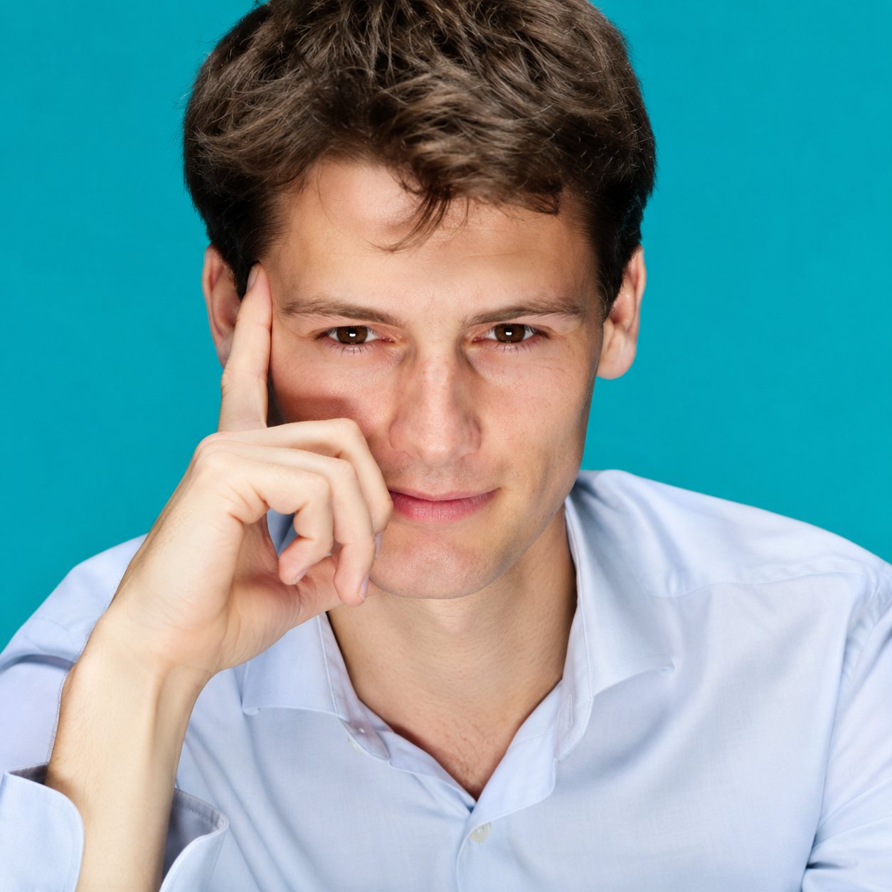 Un jeune homme aux cheveux bruns, vêtu d'une chemise bleu clair, pose le doigt sur la tempe devant un fond turquoise uni.